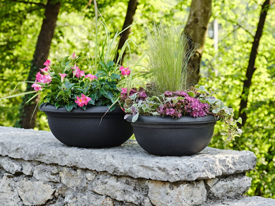 l'immagine mostra due vasi da giardino rotondo in polietilene di colore antracite, con design classico e bordo sporgente. All'interno piante fiorite.