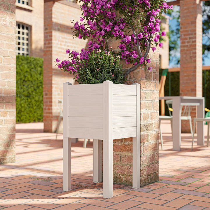 Fioriera rettangolare rialzata in plastica effetto legno chiaro con pianta rampicante viola, posizionata su pavimento in cotto all’esterno di un edificio in mattoni.