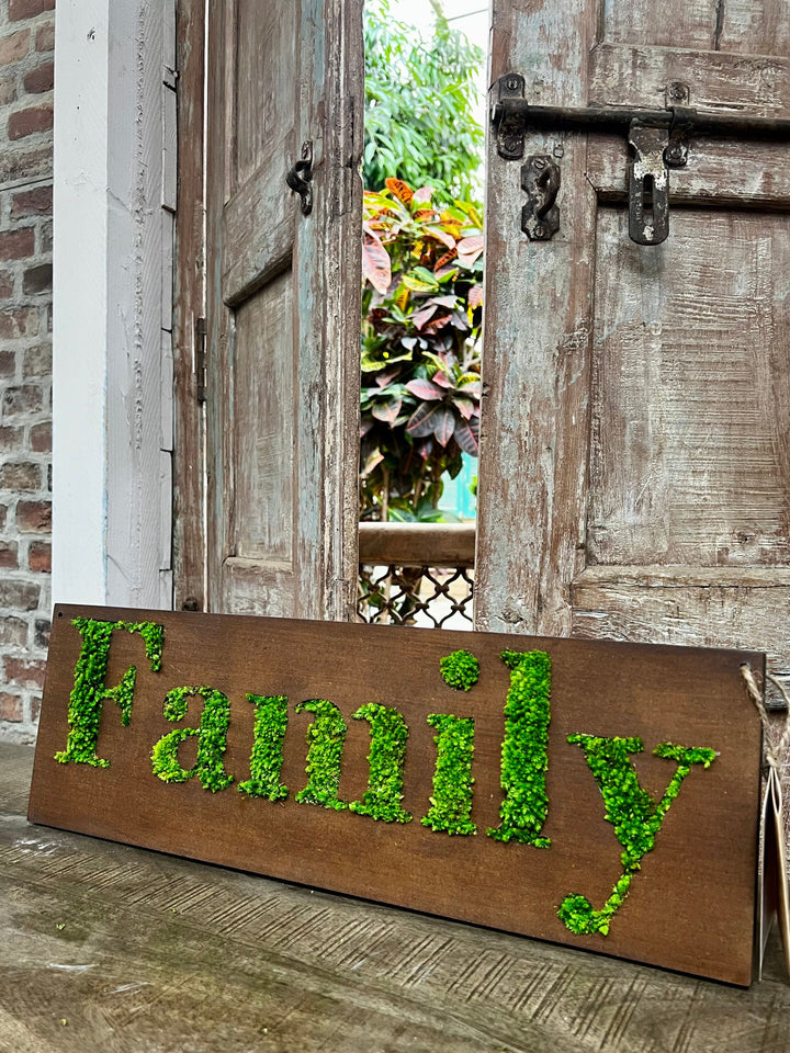 Questa immagine mostra il pannello decorativo "Family" con lettere in muschio stabilizzato posizionato in un ambiente rustico, vicino a una porta in legno antico. L'ambientazione naturale e accogliente enfatizza il design verde e organico del pannello, creando un'atmosfera calda e famigliare.