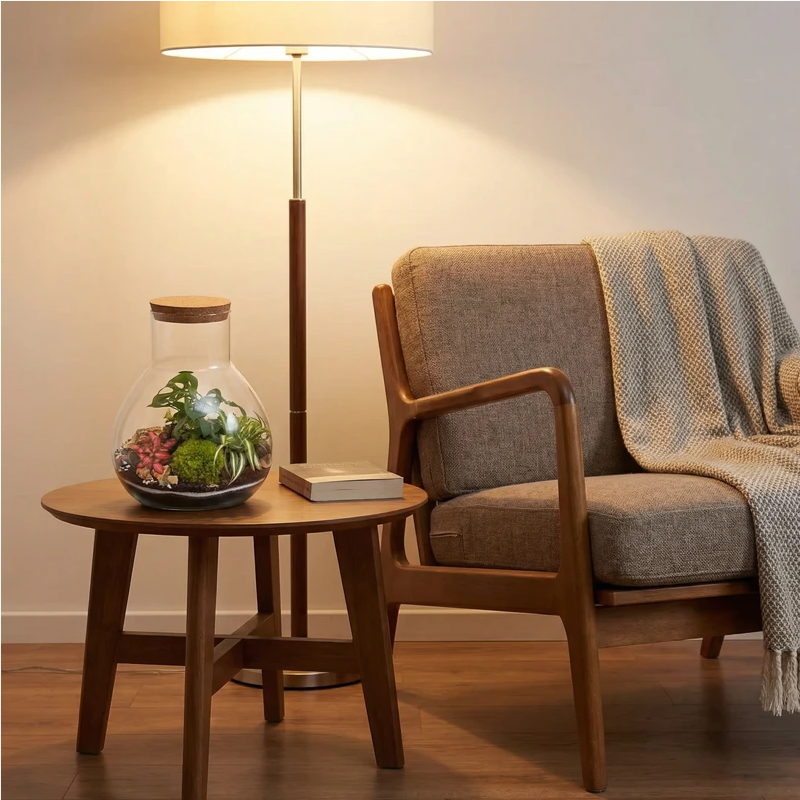 Terrarium in vetro con mini piante tropicali e muschio posizionato su tavolino accanto a poltrona, elemento decorativo naturale per soggiorno