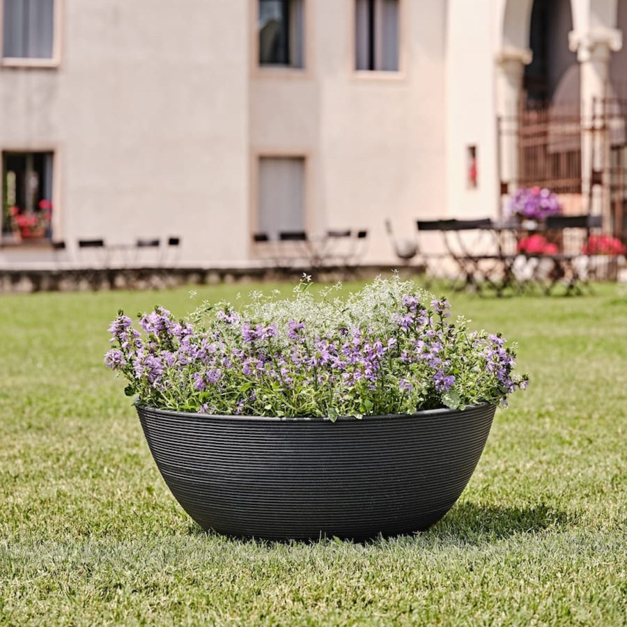 Fioriera rotonda nera con fiori lilla e bianchi posizionata su prato verde in contesto elegante.