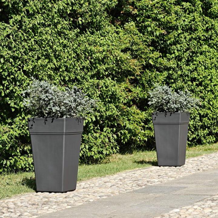Due fioriere alte antracite con piante grigio-verdi, posizionate su un vialetto lastricato accanto a una siepe verde.