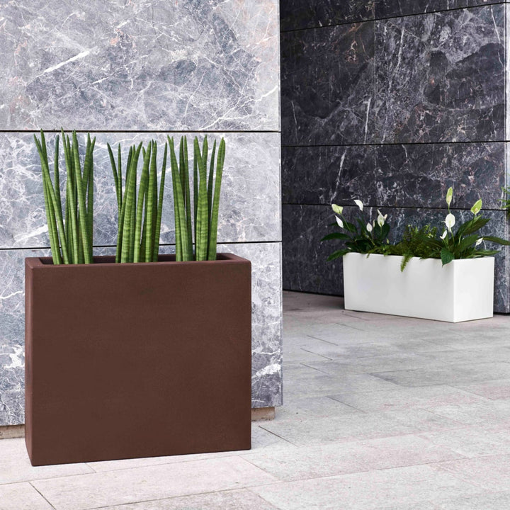 Due fioriere rettangolari moderne, una marrone con sansevieria cilindrica e una bianca con spatifillo e felci, posizionate su pavimento in pietra davanti a una parete in marmo.