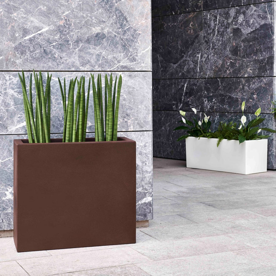 Due fioriere rettangolari moderne, una marrone con sansevieria cilindrica e una bianca con spatifillo e felci, posizionate su pavimento in pietra davanti a una parete in marmo.