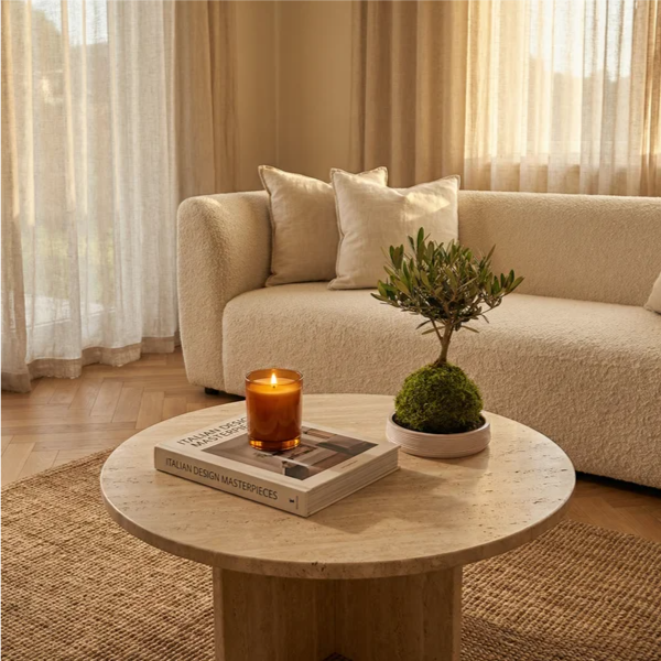  Il kokedama con la sfera di muschio verde sulla base ceramica a righe, accanto a candela ambrata accesa e libro "Italian Design Masterpieces", divano bouclé crema, golden hour