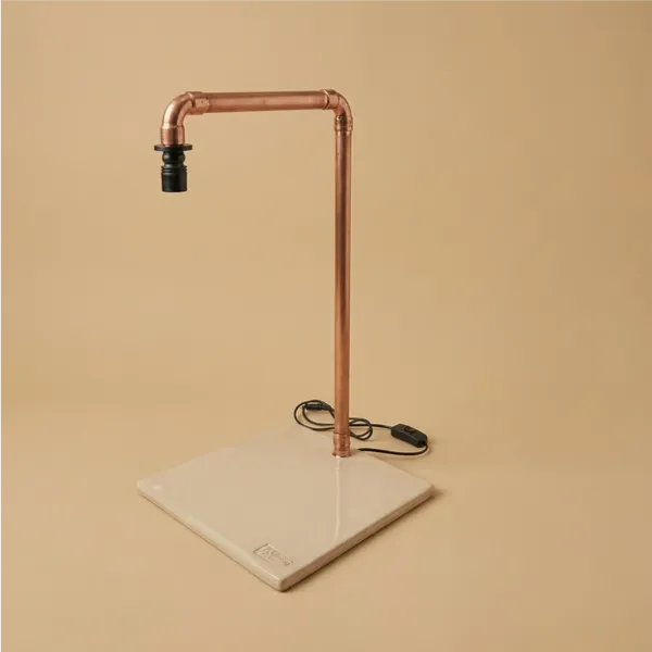 Lampada LED da tavolo industriale in tubo di rame con base quadrata in ceramica artigianale color crema, interruttore a filo nero su sfondo beige