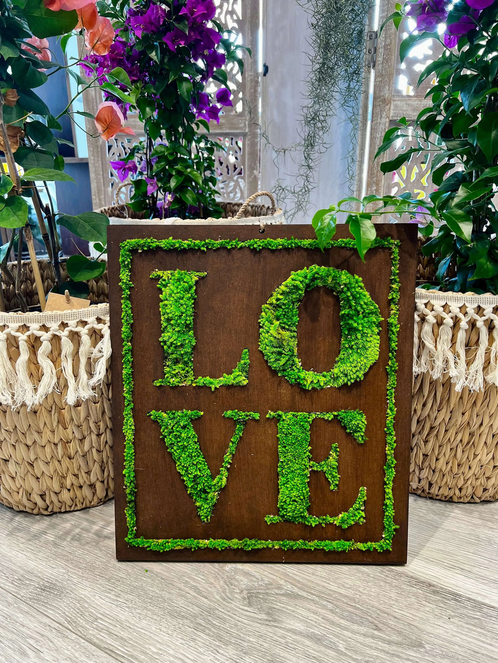 Quadro "LOVE" con scritta in muschio stabilizzato su base in legno scuro, ambientato tra piante e fiori colorati. Design quadrato 40x35 cm, decorazione naturale perfetta per interni dal tocco green ed elegante.