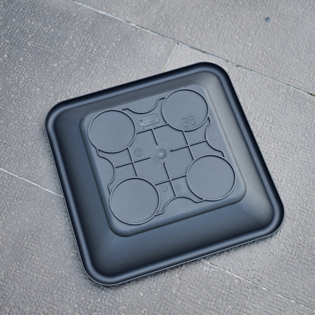 Sottovaso quadrato nero in plastica con fondo antiscivolo, visto dal basso su pavimento esterno.