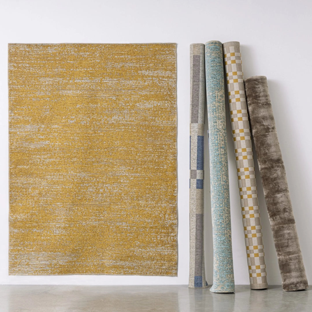 Tappeti moderni in varie fantasie e texture, esposti arrotolati accanto a un tappeto steso color senape con effetto sfumato, su sfondo neutro.