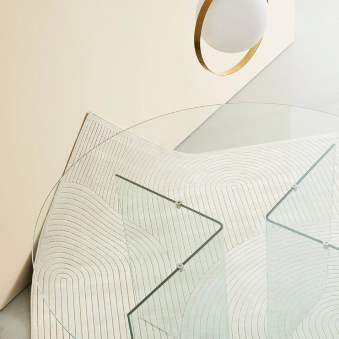 Tappeto avorio moderno con motivi curvi sotto un tavolo rotondo in vetro, in ambiente elegante e minimale.