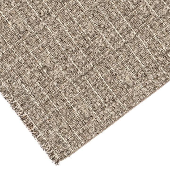 Dettaglio angolare del tappeto in fibra naturale intrecciata con disegno a quadri beige e panna e frange sui bordi.