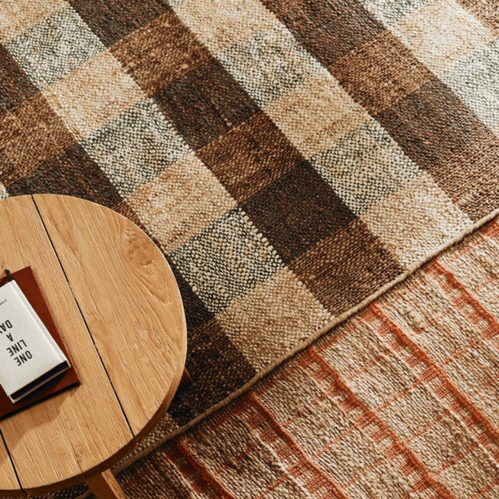 Dettaglio ambientato di tappeto in juta con motivo a scacchi nei toni del marrone, beige ed ecrù, abbinato a tavolino in legno chiaro e secondo tappeto con pattern arancione.


