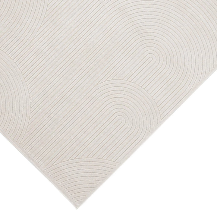 Dettaglio dell’angolo del tappeto moderno avorio con rilievo geometrico a righe curve, su sfondo chiaro.