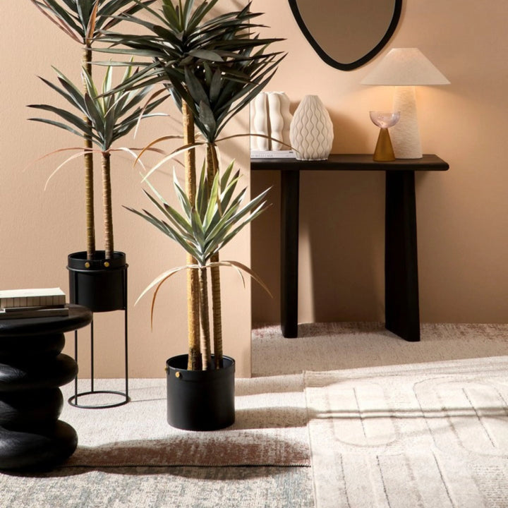 Tappeto moderno beige con effetto consumato e motivi geometrici, inserito in un angolo d’arredo con piante in vaso nero, tavolino scultoreo e consolle in legno scuro con decorazioni.

