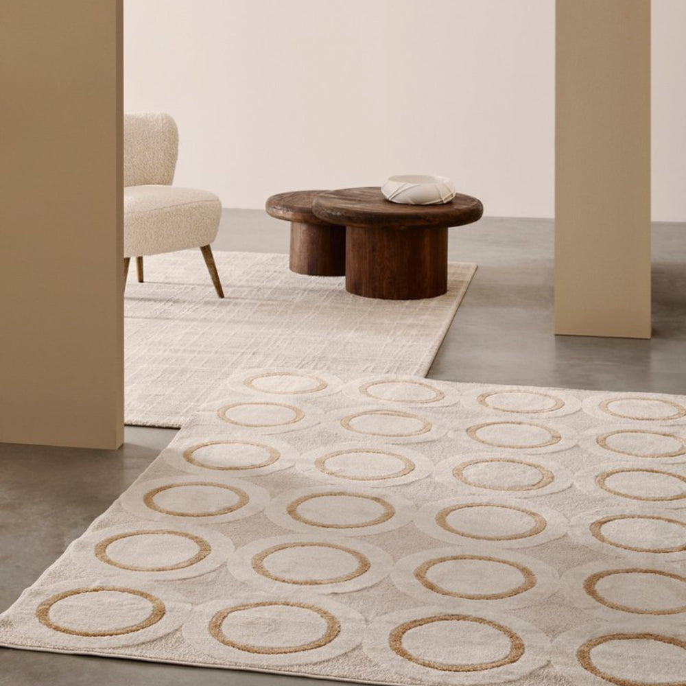 Tappeto moderno rettangolare con motivo geometrico a cerchi beige su fondo chiaro, inserito in un ambiente minimal con tavolino in legno e poltrona in tessuto bouclé.