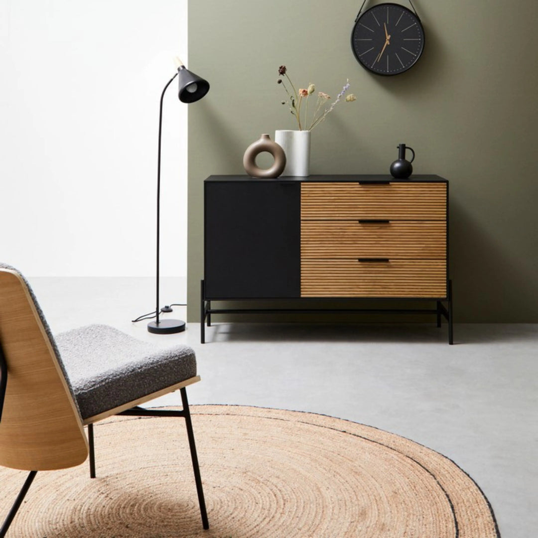Tappeto rotondo in juta naturale con bordo nero in un salotto dal design scandinavo, con sedia in legno e tessuto bouclé, madia moderna nera e legno, e lampada da terra minimal.

