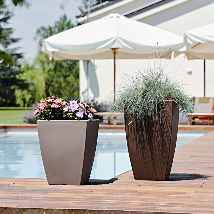 Coppia di vasi alti da esterno posizionati a bordo piscina, contenenti fiori rosa e erba ornamentale, ideali per decorare ambienti outdoor moderni.

