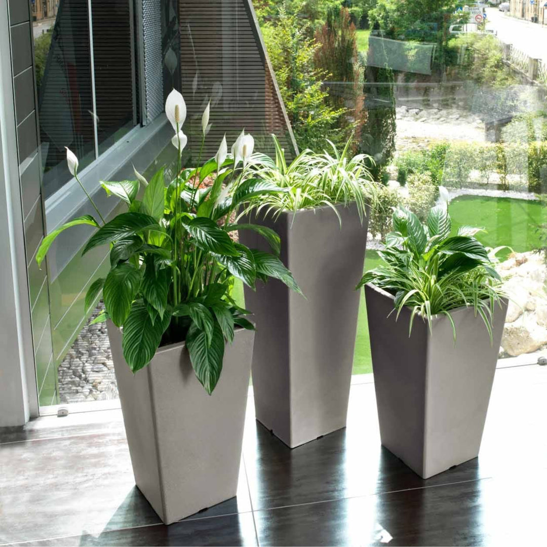 Vasi alti moderni in finitura satinata con piante verdi e spatifillo, posizionati all’interno di uno spazio luminoso e contemporaneo con vista sul giardino