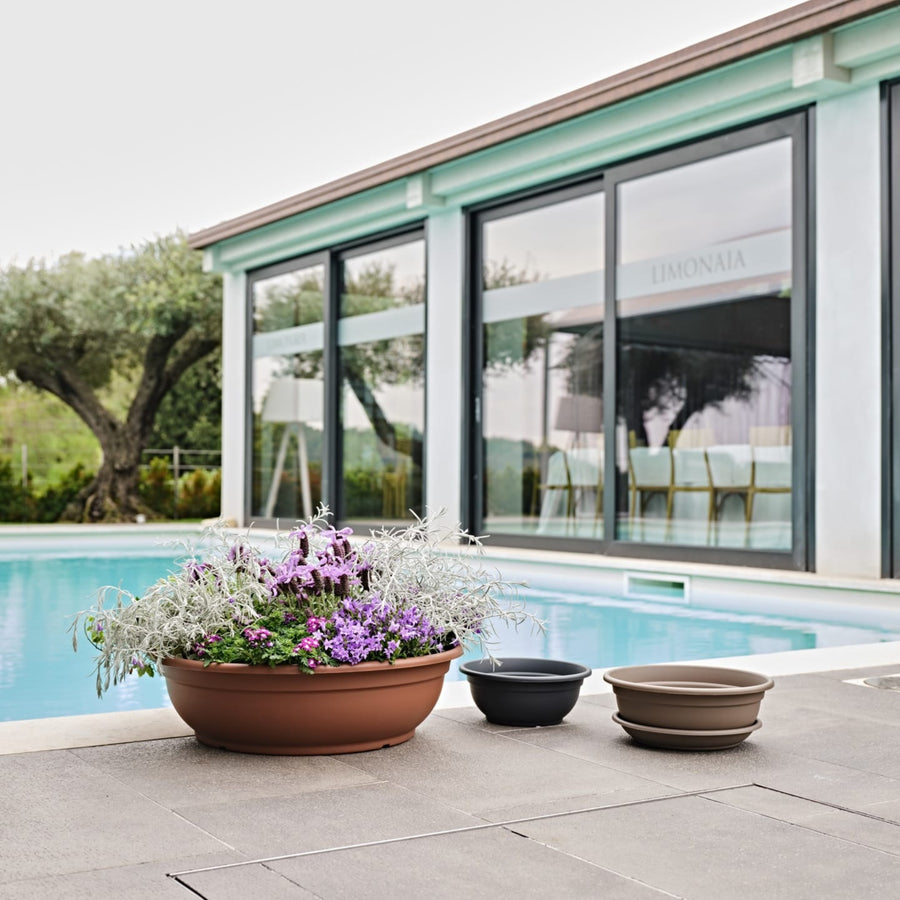 Vasi da giardino bassi color terracotta, antracite e tortora con fiori stagionali viola e grigi, posizionati a bordo piscina in un ambiente moderno.